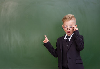 Surprised boy stands near empty chalkboard and showing finger up