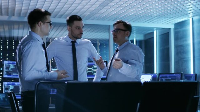 Three Technical Moderators Have Active Discussion in Monitoring Room. System Control Room is full of Working Displays Showing Various Data and Has Servers Racks. 