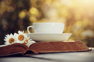 White cup, bouquet of camomiles and old open book.