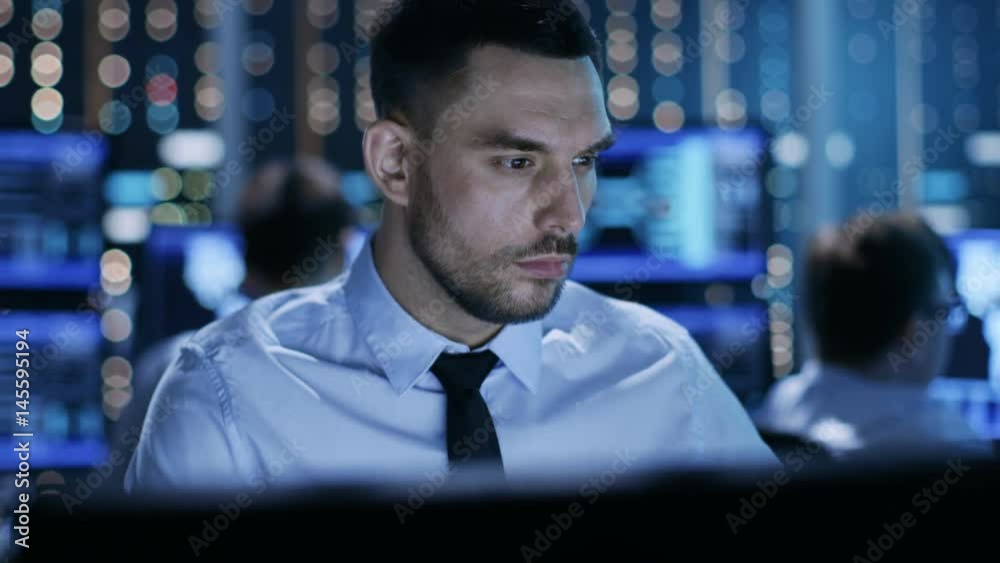 Poster  Close-up of a Professional Technical Controller Sitting at His Desk with Multiple Displays Before Him. In the Background His Colleagues Working in System Control Center. 