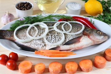 Raw fish lies on a platter among vegetables and greens before cooking.