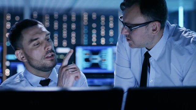 Two Professional Technicians Discussing Problems Shown on Multiple Monitors in a System Control Center. Shot on RED EPIC-W 8K Helium Cinema Camera.