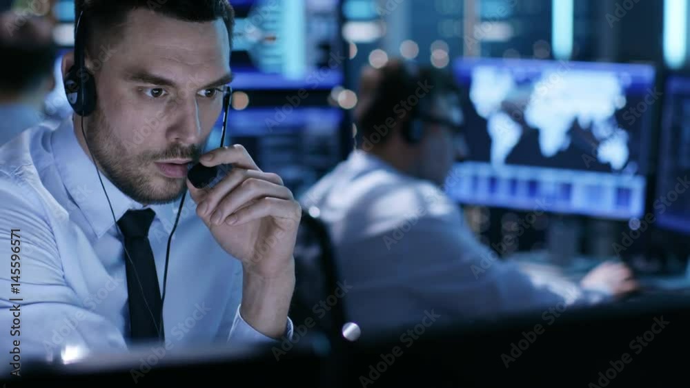 Poster in monitoring room technical support specialist speaks into headset. his colleagues are working in t