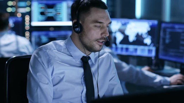 In System Control Center Technical Support Specialist Speaks into Headset. His Colleagues are Working in the Background in a Room Full of Screens.  Shot on RED EPIC-W 8K Helium Cinema Camera.