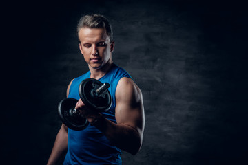 A man dressed in a blue sportswear holds the dumbbell over dark grey background.