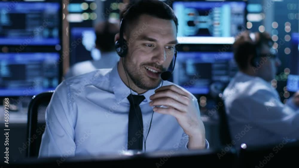 Poster in system control center technical support specialist speaks into headset. his colleagues are workin