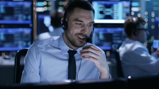 In System Control Center Technical Support Specialist Speaks into Headset. His Colleagues are Working in the Background in a Room Full of Displays.  Shot on RED EPIC-W 8K Helium Cinema Camera.