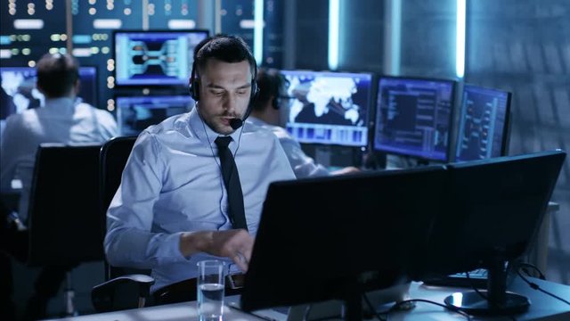 In System Control Center Technical Support Specialist Speaks into Headset while Sitting at His Desk Before Multiple Monitors. His Colleagues are Working in the Background in a Room Full of Displays. 