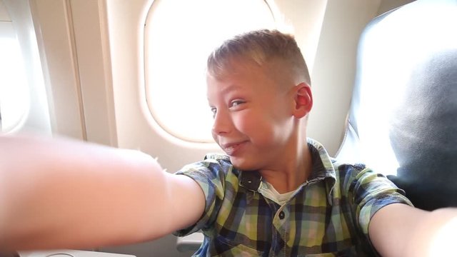 Closeup portrait of cute funny kid taking selfie in plane, makes silly faces and gives thumb up. Happy child sits on his seat near window with bright sunlight through it in background. Wide angle.