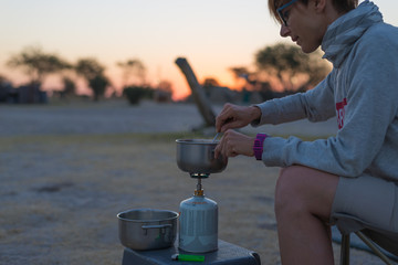 Woman cooking with gas stove in camping site at dusk. Gas burner, pot and smoke from boiling water. Adventures in african national parks.
