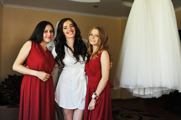 Happy cheerful brunette bride wearing at her room with two bridesmaids on red dress.