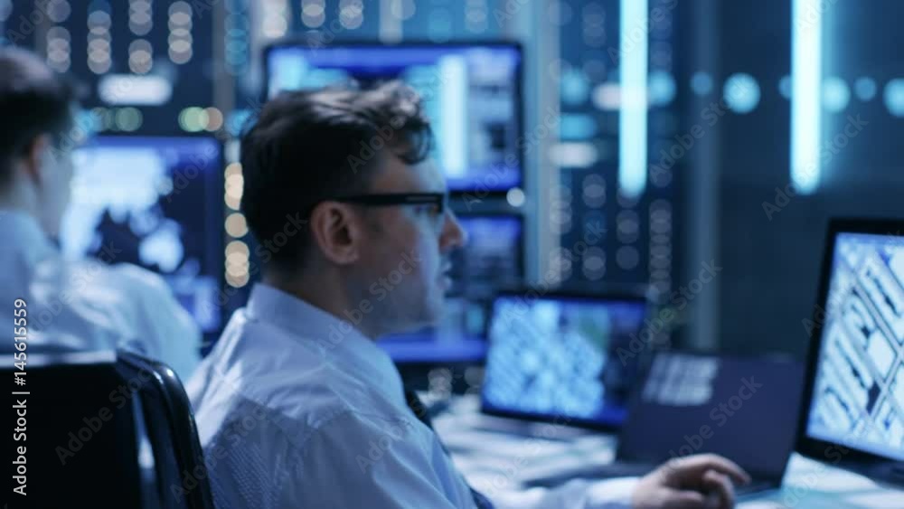 Poster In the Monitoring Room Team of System Engineers Working at Their Workstations. Multiple Displays Show Various Important Data and Information. Possible Air Traffic/ Power Plant/ Security Room Theme. 