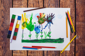 Child draws a pencil drawing of the flowers on green grass and blue sky on wood background. Top view