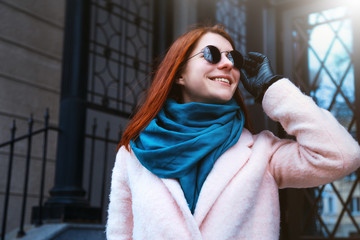 Red haired beautiful girl is walking by the street in a pink coat and blue scarf, with sunglasses.