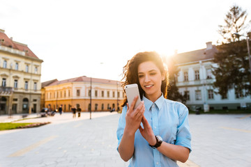 Beautiful young woman is using an app in her smartphone device to send a text message in front of a...