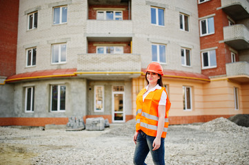 Obraz na płótnie Canvas Engineer builder woman in uniform waistcoat and orange protective helmet against new building. Property living block theme.