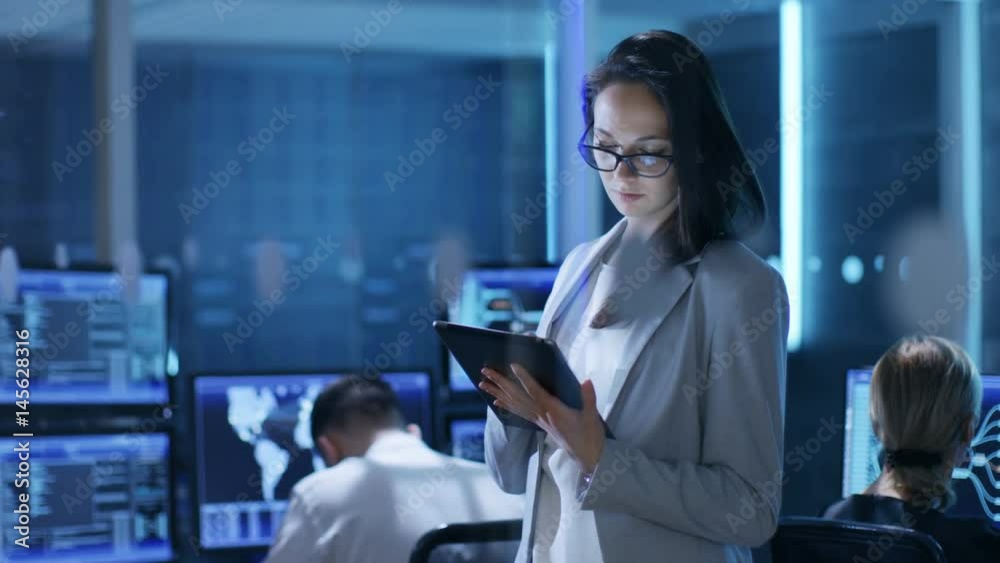 Aufkleber young female engineer uses tablet in system control center. in the background her coworkers are at t