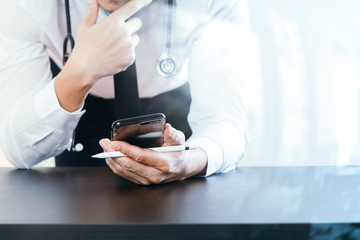close up of smart medical doctor working with smart phone and stethoscope on dark wooden desk with glass reflected view