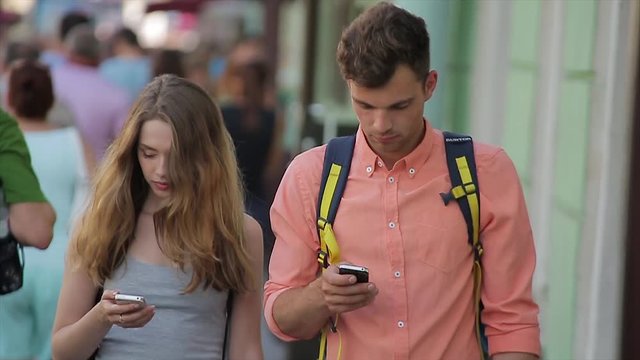 Young couple walk along the busy city street and look at their mobiles in slo-mo