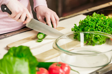 female hands with a knife sliced fresh cucumber on a wooden board. On the table in the kitchen are vegetables.