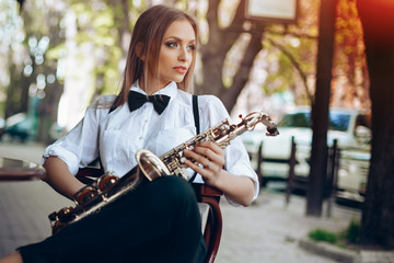 Young attractive girl in white shirt with a saxophone sitting near coffee shop - outdoor in street....