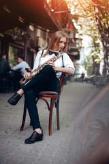 Playing young attractive girl in white shirt with a saxophone sitting near coffee shop - outdoor in street. Sexy young woman with sax playing melody