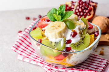 Fresh fruit salad with yogurt and walnuts in glass bowl on stone background.
