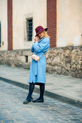 Fototapeta na wymiar Young beautiful girl with perfect smile in a blue coat and burgundy hat walking down the street and talking on mobile phone