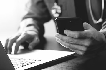 close up of smart medical doctor working with mobile phone and laptop computer and stethoscope on dark wooden desk,black and white