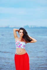 Portrait of young beautiful woman wearing a t-shirt and red skirt at the hips, with piercings in the navel. Girl on the beach straightens flowing hair.