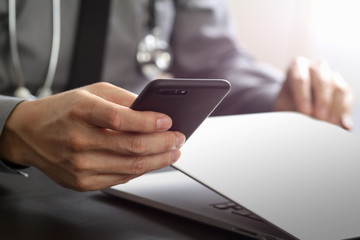 close up of smart medical doctor working with mobile phone and laptop computer and stethoscope on dark wooden desk