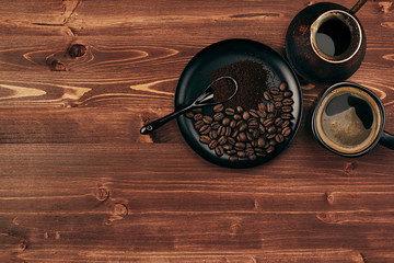 Hot coffee in black cup with beans, spoon and turkish pot cezve with copy space on brown old wooden board background, top view.  Rustic style.