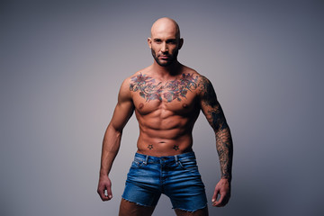 Shaved head, muscular male with tattoos on his torso over grey vignette background.