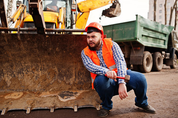 Fototapeta na wymiar Brutal beard worker man suit construction worker in safety orange helmet, against traktor with hammer at hand.