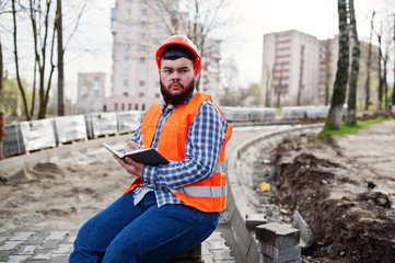 Brutal beard worker man suit construction worker in safety orange helmet sitting on pavement and makes working notebook entries.