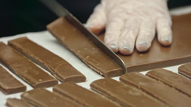 Closeup of female hands in protective gloves cutting chocolate fudge with knife into bars on table