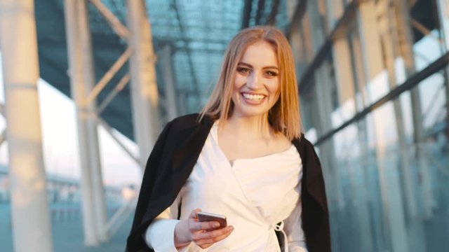 Beautiful blonde European girl walking through the airport terminal, using her phone, looks right towards the camera and gives a happy smile. Stylish wear, elegant white blouse, red tight trousers.