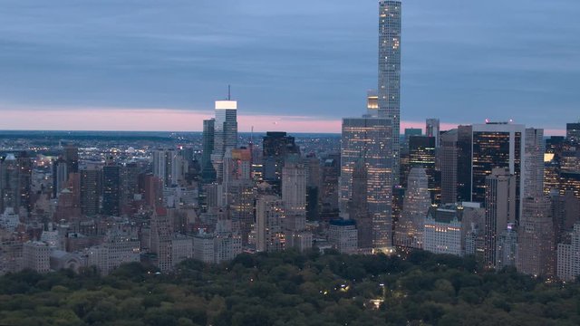 AERIAL HELI SHOT: Lights shining in windows of giant glassy skyscrapers, office buildings and luxury condominiums overlooking lush green Central park on tranquil early morning, New York City Manhattan