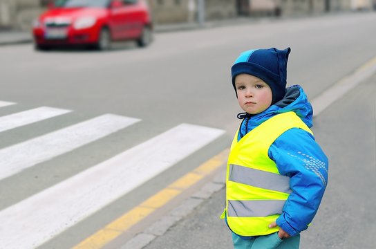 Child in front of pedestrian crossing. Little boy finds out if he can cross the crossswalk. He wears reflective vest because of safety. Car in the background. Child concept. Traffic concept.