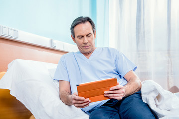 middle aged patient using digital tablet and sitting on bed in hospital