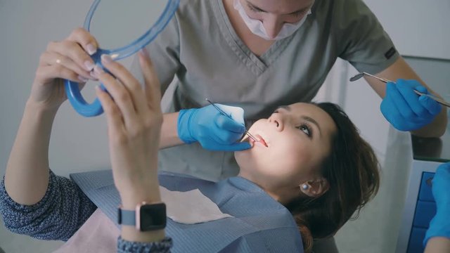 Woman visiting the dentist. Female lying on dental chair and holding the mirror. Orthodontist checks-up the overbite.