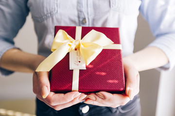 Close up shot of female hands holding a red gift wrapped with yellow ribbon. A present in the hands of a woman indoor. Female gives a gift to another person.