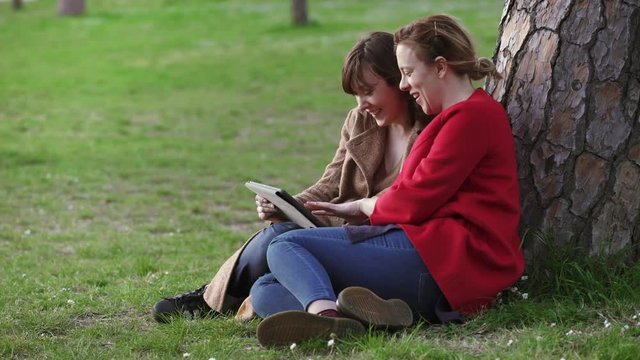 Two relaxed friends sitting under a tree use the tablet