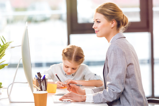 Young Mother Working With Computer While Her Daughter Drawing In Business Office