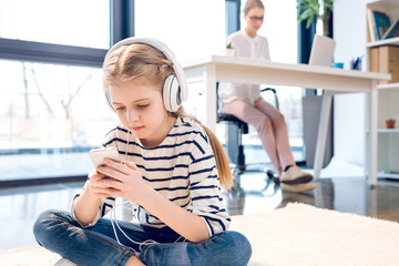 adorable daughter using smartphone and headphones, businesswoman working behind in office