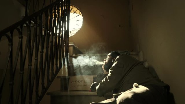 Homeless smokes a cigarette looking at the light, lying on the stairs of buildin