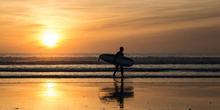 Silhouette of Surfer with surfboard walking out of the ocean during sunset