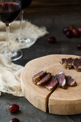 Slices of dried meat on wooden cut, red grapes and two glasses of red wine on dark rustic background