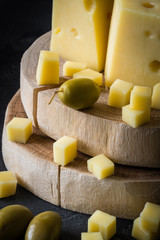 Close up of Swedish hard yellow cheese with holes chopped on wooden slices with green olives on dark rustic background