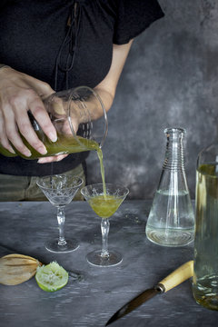 A women is pouring a matcha wine cocktail. Photographed from front view on a grey background.